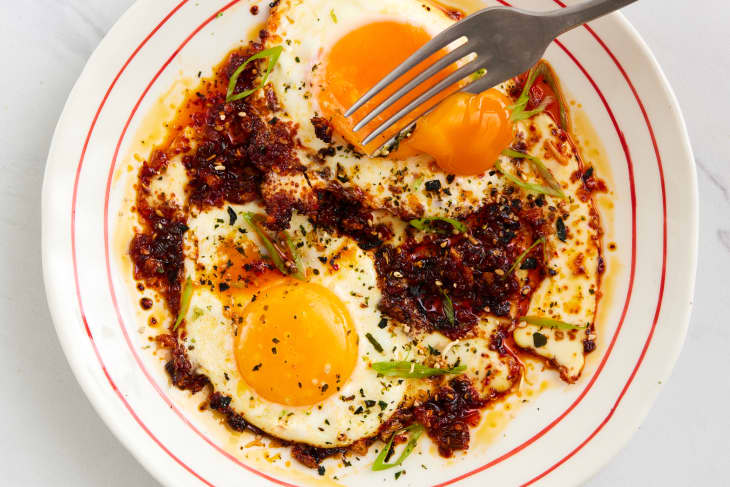 two sunny side up eggs with chili crisp on a white plate with red concentric circles, garnished with sesame seeds and scallions with a fork breaking one of the yolks