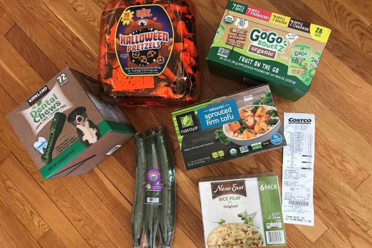 Assorted grocery items on wooden floor, including Halloween pretzels, GoGo squeeZ, tofu, cucumbers, rice pilaf, and dental chews.