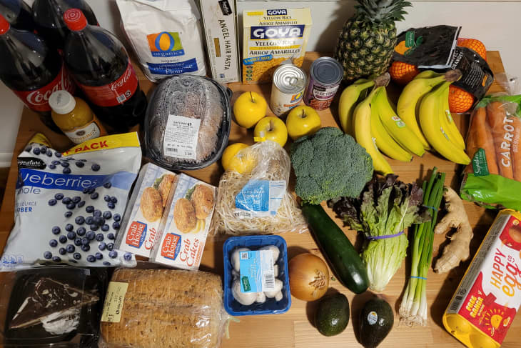 Assorted groceries on a table, including bananas, broccoli, pineapple, eggs, bread, blueberries, and a rotisserie chicken.