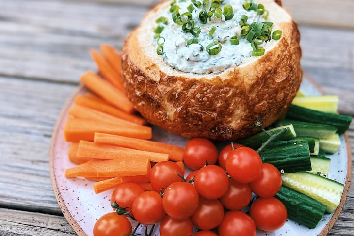 spinach dip in a bread bowl surrounded by cherry tomatoes, chopped carrots, cucumbers