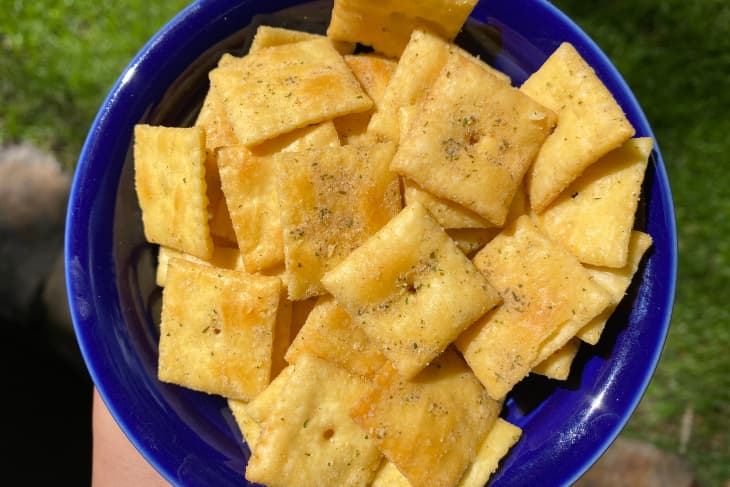 Bowl filled with seasoned square crackers, held against a green grass background.