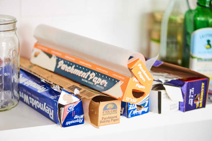 Parchment paper and aluminum foil boxes on a kitchen shelf next to a glass jar and bottles.