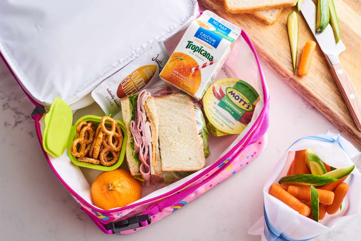 Pink lunchbox with a ham sandwich, pretzels, orange, applesauce, juice box, and almond butter packet.