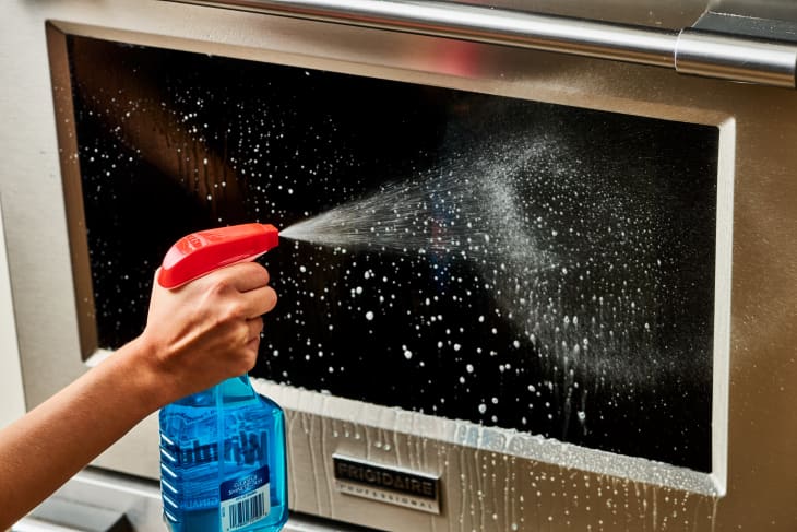 A person spraying glass cleaner onto the glass of their stainless steel oven's door