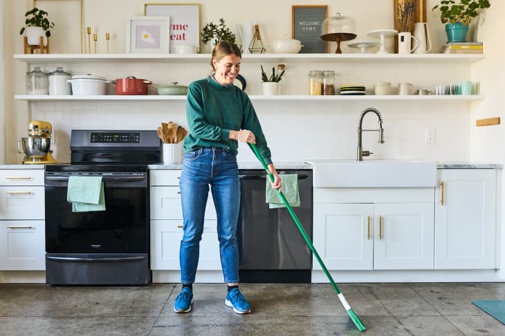 A woman standing in a white kitchen, sweeping the concrete floors with a broom