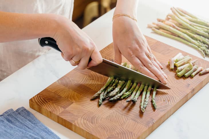 Someone cutting asparagus on wooden cutting board with chef's knife.