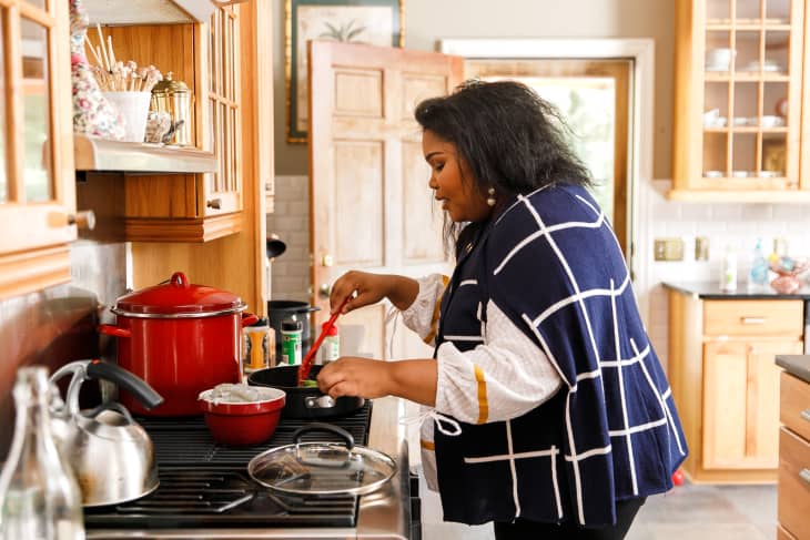 Woman cooking in a kitchen with wooden cabinets, using a red pot on the stove.