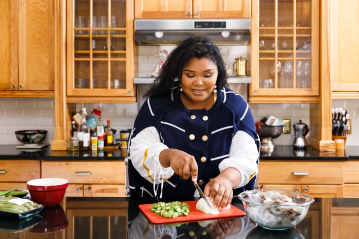 Woman chopping vegetables in a kitchen with wooden cabinets and various cooking ingredients on the counter.