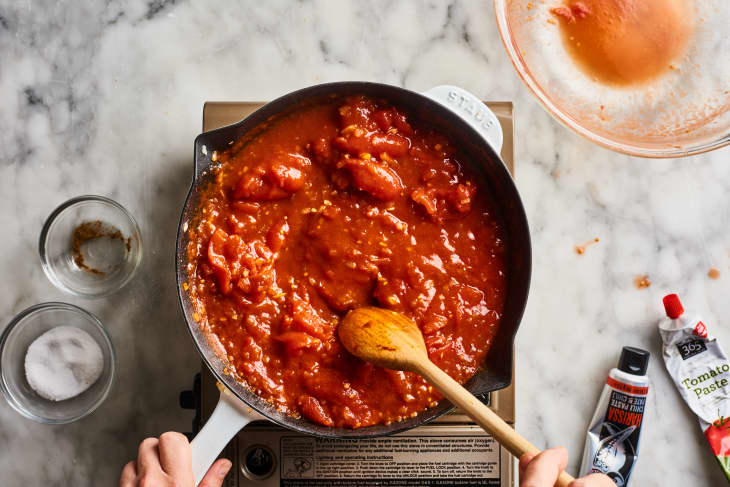 Cooking tomato sauce in a pan on a marble countertop with wooden spoon, tomato paste, and spices nearby.