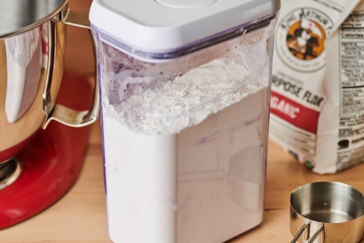 Flour in plastic container with bag in the background, measuring cup, stand mixer