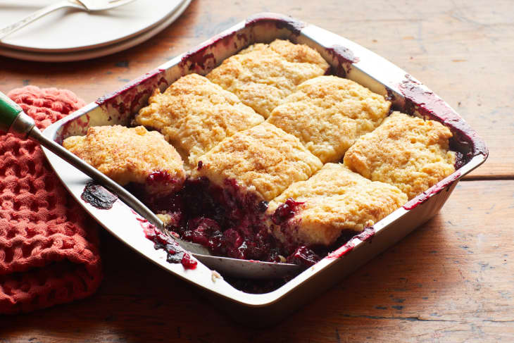 The cooked berry cobbler sits on a table. A spoon rest inside the pan after many pieces have been removed.