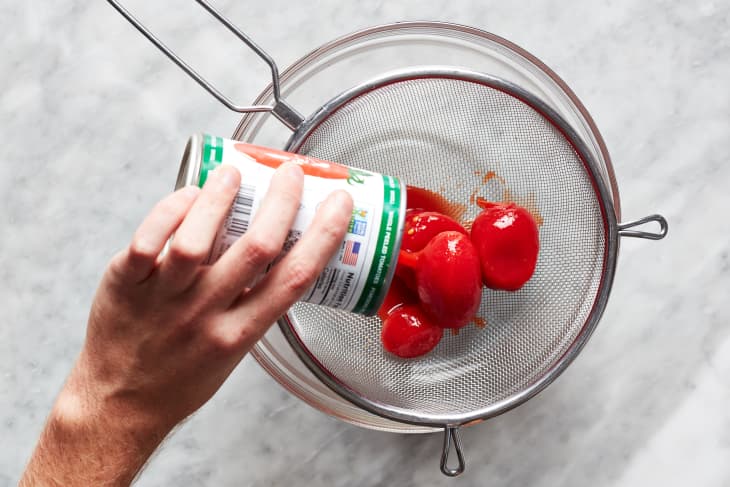 Canned tomatoes being poured into strainer over glass bowl.