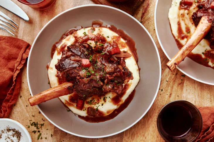 A braised lamb shank sits in a circular plate surrounded by a napkin, plates of garnish, and utensils.