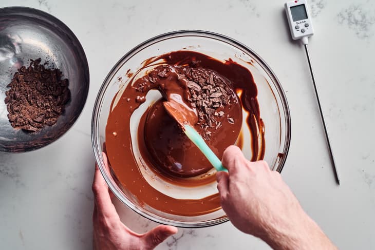Melted chocolate being stirred in a glass bowl with chocolate shavings and a thermometer nearby.