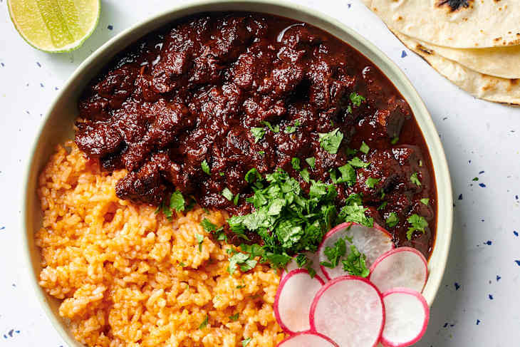 Overhead view of chile colorado and rice in a green bowl topped with radishes and cilantro. Half a lime in the top left corner of the shot, and 3 tortillas peeking in on the top right corner of the shot.