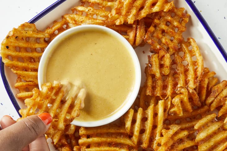 Overhead shot of waffle fries, and a small white bowl of chick fil a sauce on a white dish with a blue rim. In the bottom left of the image theres a hand dipping a waffle fry into the sauce.