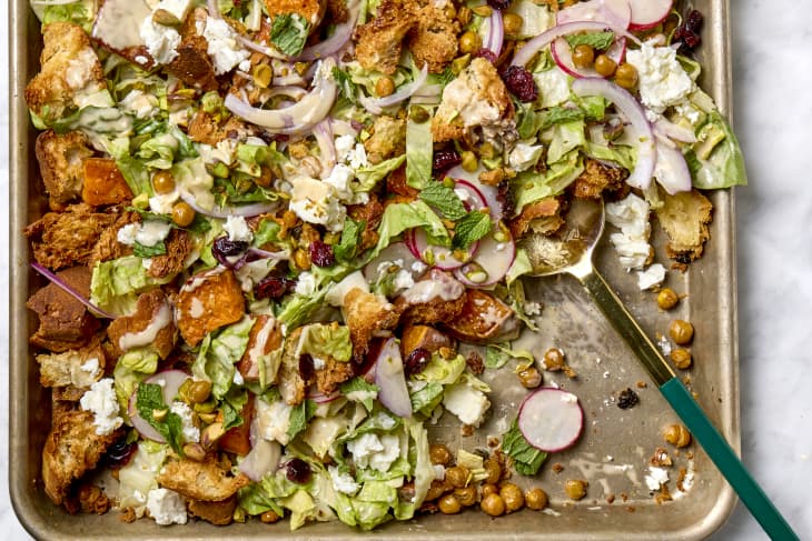 Overhead shot of a sheet pan salad with sweet potatoes, red onion, radishes, chickpeas and feta, and a gold and green serving spoon resting in the pan.