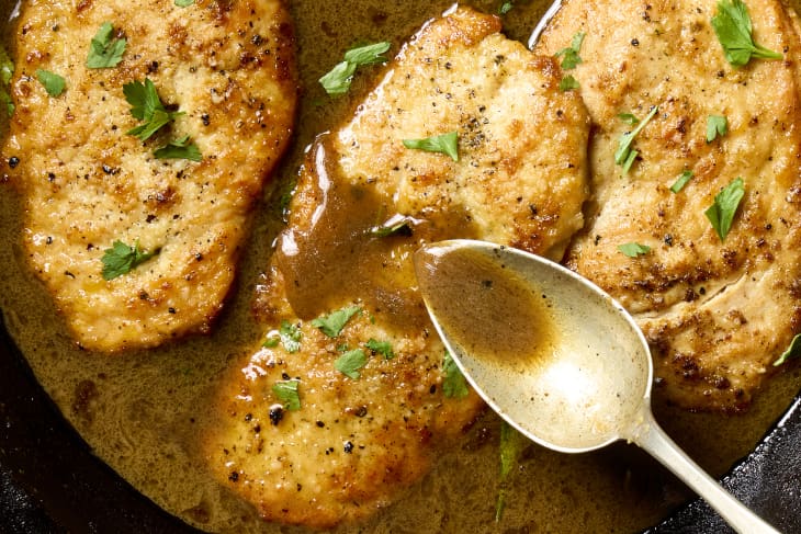 close up shot of chicken scallopini in a cast iron pan, topped with herbs, and a spoon drizzling sauce from the pan over the chicken.