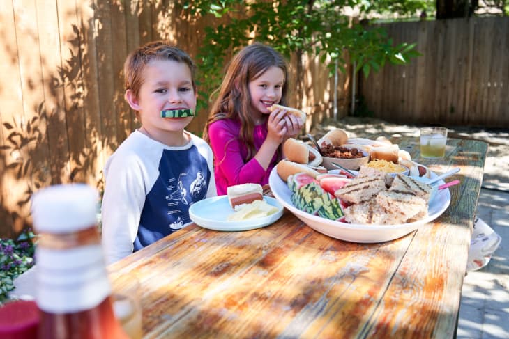 A young boy and girl sit at a picnic table eating hot dogs and watermelon from the hot dog snack board.