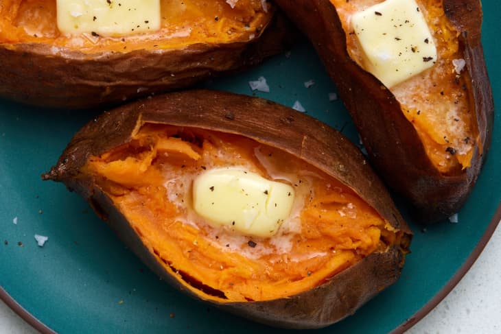 Overhead view of 3 baked sweet potatoes cut open on a dark blue plate, topped with butter and black pepper.