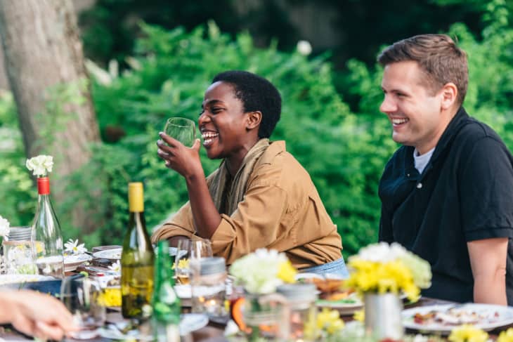Two people laughing at an outdoor table with wine bottles and flowers.