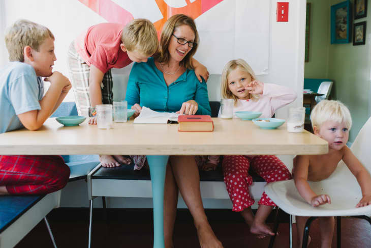 Family gathered around a dining table, mother reading a book, children in pajamas, with bowls and glasses on the table.