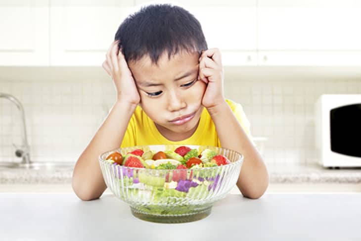 Child in yellow shirt frowning at a bowl of mixed salad with tomatoes, lettuce, and cucumbers in a kitchen setting.