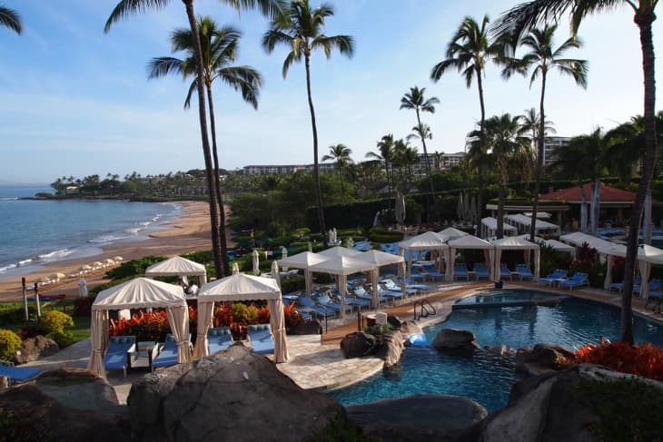 Beachfront resort with palm trees, cabanas, and a pool overlooking a sandy beach and ocean.
