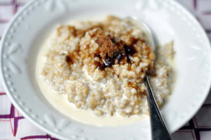 A silver spoon is placed in a white bowl of steel-cut oats soaked in milk and topped with brown sugar and syrup.