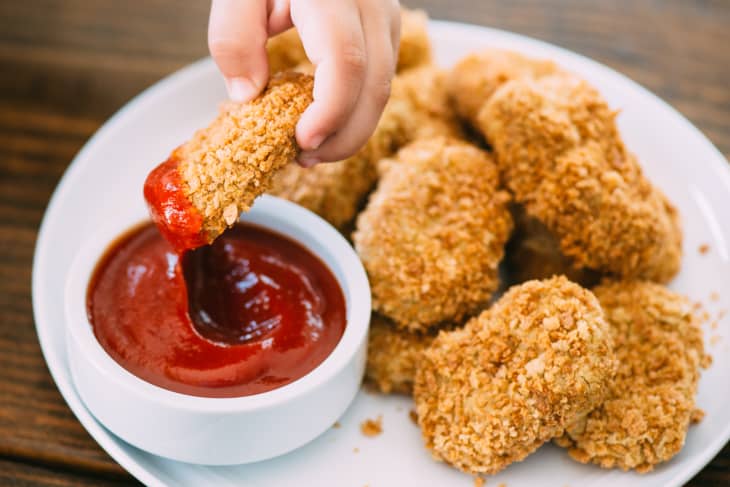 Hand dipping a crispy chicken nugget into a bowl of ketchup on a white plate.
