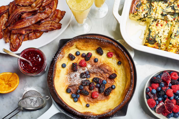 A table set for brunch with a Dutch baby, bacon, breakfast casserole, berries, jam, and mimosas