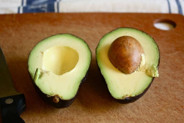 Halved avocado on a wooden cutting board with a knife nearby.
