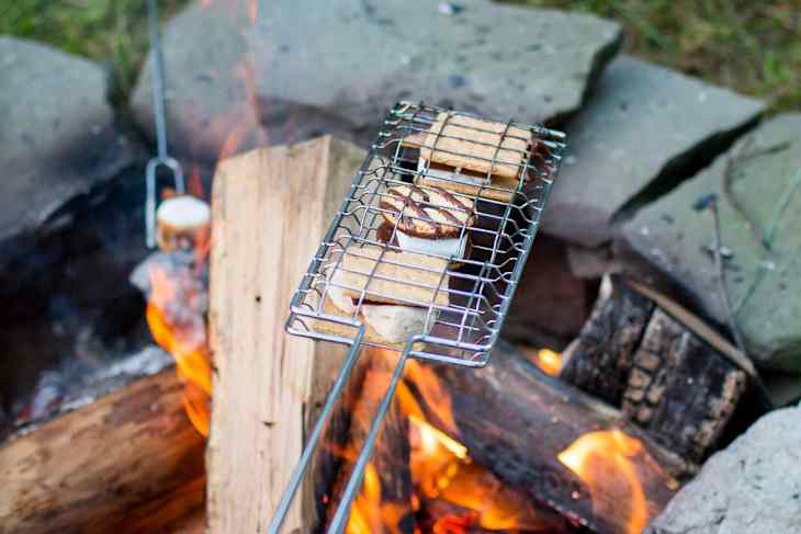 Person roasting marshmallows over a campfire with a grill basket, surrounded by stone fire pit and grass.