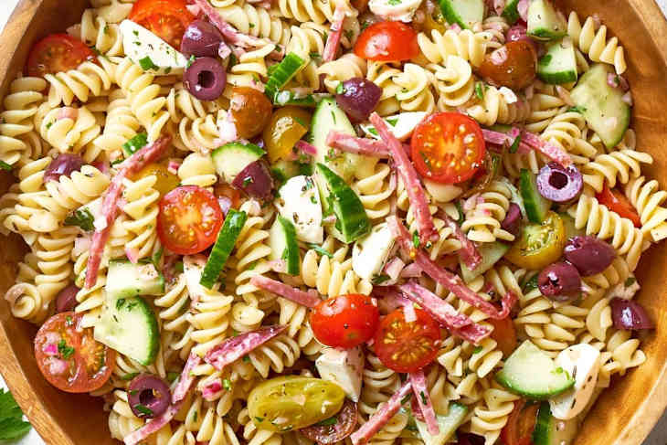 Pasta salad with cherry tomatoes, chopped cucumbers, red onions, olives, and other ingredients in a wooden bowl
