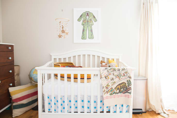 White crib with star-patterned bedding, a colorful blanket, stuffed animals, and a framed green suit illustration above.