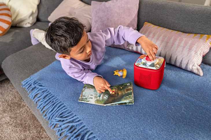 A child in a purple shirt plays on a blue blanket, interacting with a red toy speaker and a storybook.