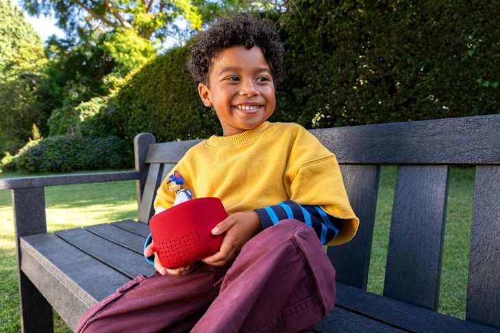 A smiling child in a yellow shirt and striped sleeves sits on a wooden bench, holding a red speaker with a toy figure.