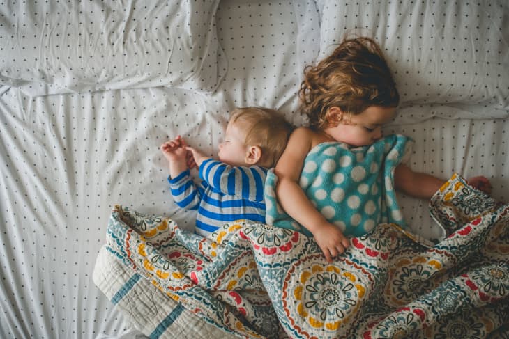 Two children sleeping on a bed, one in blue stripes, the other with a polka dot blanket, under a colorful quilt.