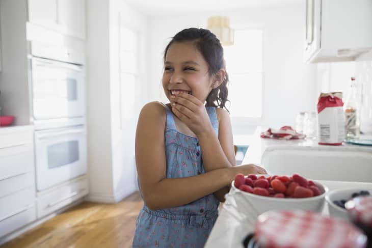Girl in a blue dress smiling in a bright kitchen with a bowl of strawberries on the counter.
