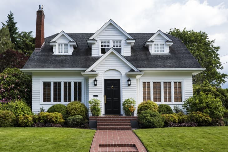 White two-story house with black door, dormer windows, and manicured shrubs in front yard.