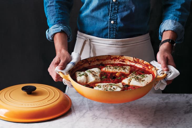 Someone holding Le Creuset cast iron braiser with braised fish in tomato sauce.
