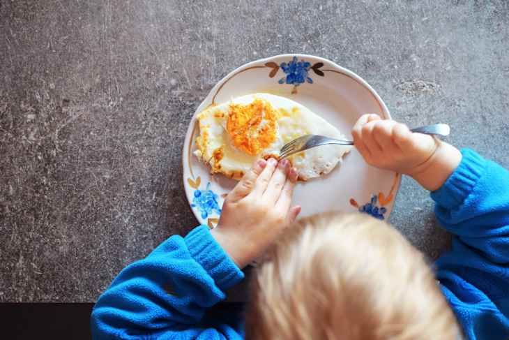 Two years old kid boy eats fried egg on white flower patterned plate on grey grunge table background. Kid learns to use tableware.