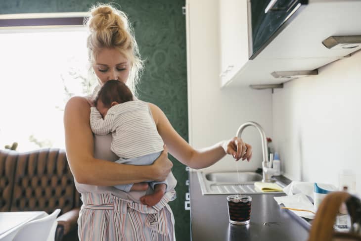 Mother holding newborn baby in kitchen while making tea