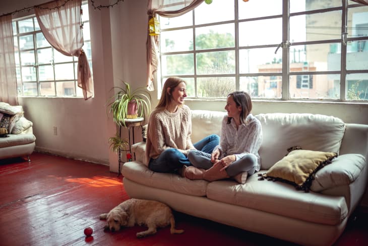 mother and teen daughter talking on sofa, dog on floor in front of them, large windows behind them
