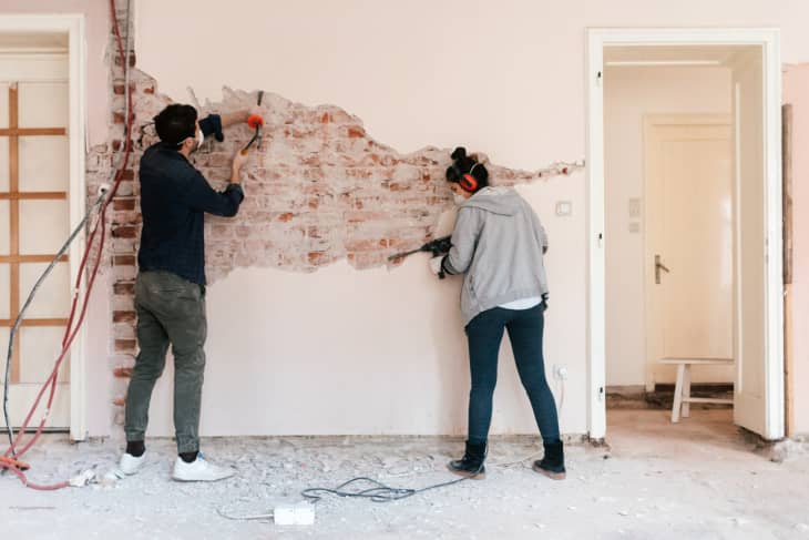 Two people removing plaster from a brick wall in a room under renovation.