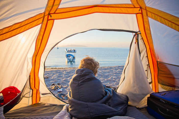 A child wrapped in a sleeping bag sits inside a tent, looking out at a beach and a boat on the water.