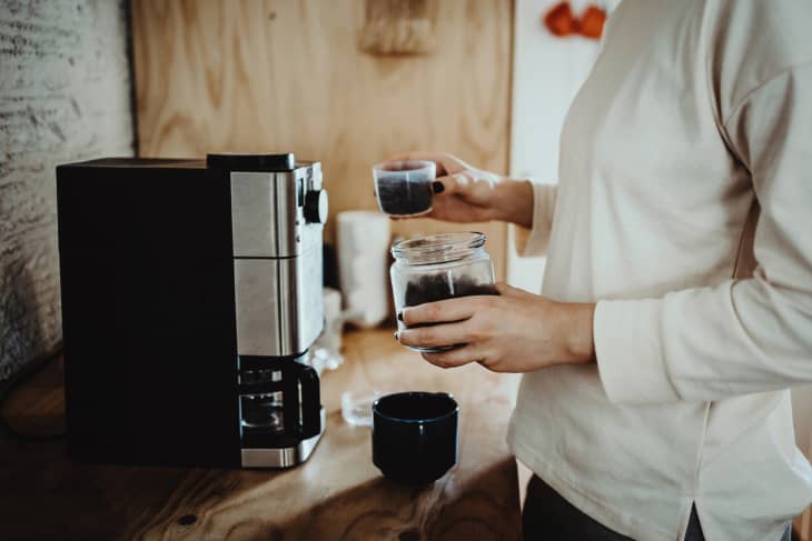 Young woman measuring and pouring roasted coffee beans into coffee machine and preparing coffee at home in the early morning.