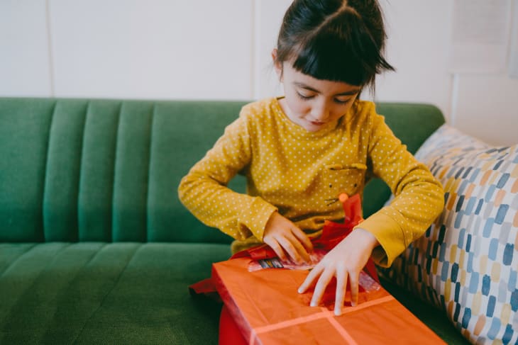 Little girl wearing yellow long sleeved shirt opening red-paper-wrapped gift on green velvet sofa. She is concentrating.