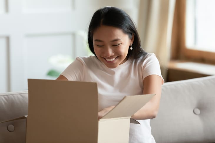 Woman sitting on sofa in living room; opens received delivered parcel and feels happy.