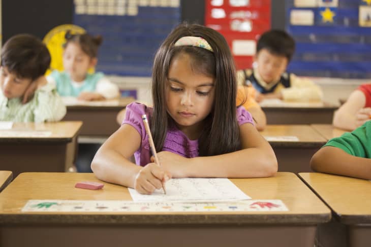 girl sitting at desk writing, wood desk, kids classroom, headband, pencil, worksheet